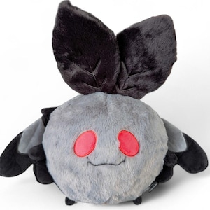 Puede incluir: Un juguete de peluche redondo con forma de polilla, cuerpo gris difuso, dos grandes antenas negras en la parte superior y alas negras m&aacute;s peque&ntilde;as a los lados. La criatura tiene dos ojos ovalados de color rojo brillante y una boca cosida.