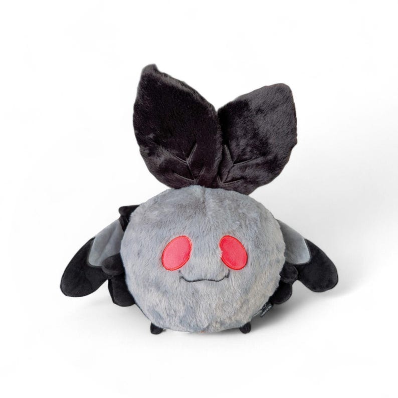 Puede incluir: Un juguete de peluche redondo con forma de polilla, cuerpo gris difuso, dos grandes antenas negras en la parte superior y alas negras m&aacute;s peque&ntilde;as a los lados. La criatura tiene dos ojos ovalados de color rojo brillante y una boca cosida.
