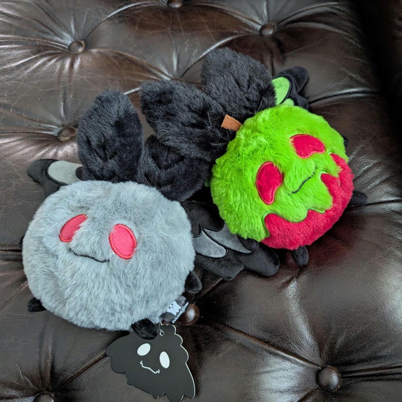 Puede incluir: Dos peluches redondos con alas negras. Uno es gris claro con ojos bordados rojos y una boca peque&ntilde;a. El otro es verde brillante con una mancha roja, un peque&ntilde;o tallo marr&oacute;n y ojos bordados. Tiene una etiqueta negra con un gr&aacute;fico de un fantasma blanco.