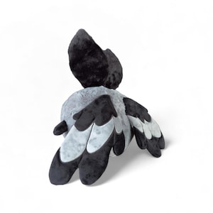 Puede incluir: Un juguete de peluche con forma de ave que presenta un cuerpo redondo de color gris claro con la cabeza y el cuello de color negro oscuro. Las alas est&aacute;n formadas por paneles de tela superpuestos en negro y gris claro, con forma redondeada.