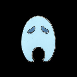Pu&ograve; includere: Spilla in smalto a forma di fantasma azzurro con bordo color oro. La spilla presenta due occhi blu a forma ovale e un'apertura curva nella parte inferiore. La spilla &egrave; posta su uno sfondo nero.