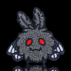 May include: Embroidered patch featuring a stylized moth-like creature with a fluffy gray body, red eyes, and black and white wing details. The patch has a smiling face and is set against a black background.