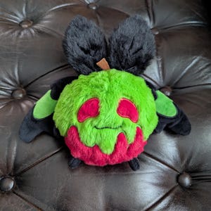 Puede incluir: Un peluche con forma de manzana verde redonda con un peque&ntilde;o tallo marr&oacute;n en la parte superior. El juguete presenta dos grandes orejas negras difusas, alas negras con detalles verdes, dos ojos ovalados rojos y un patr&oacute;n rojo que gotea en la base.