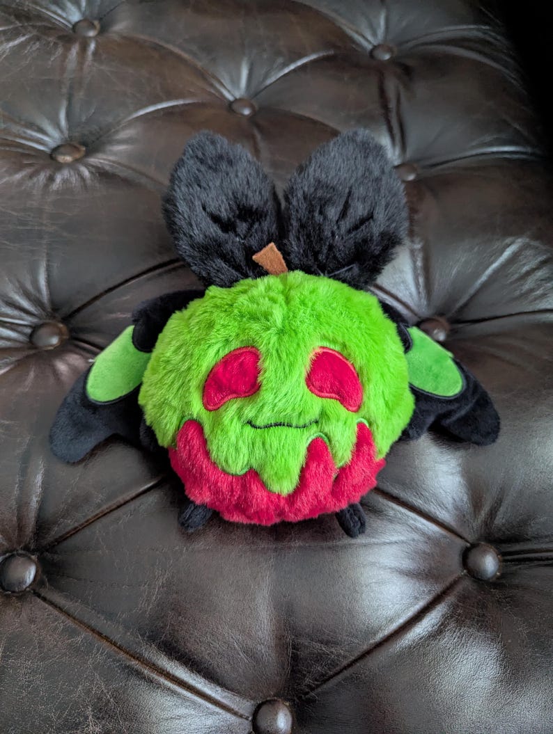 Puede incluir: Un peluche con forma de manzana verde redonda con un peque&ntilde;o tallo marr&oacute;n en la parte superior. El juguete presenta dos grandes orejas negras difusas, alas negras con detalles verdes, dos ojos ovalados rojos y un patr&oacute;n rojo que gotea en la base.