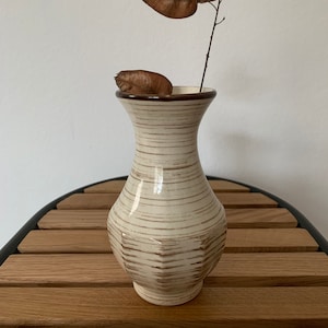May include: A white ceramic vase with brown stripes and a geometric design. The vase is filled with a single dried branch with brown seed pods.