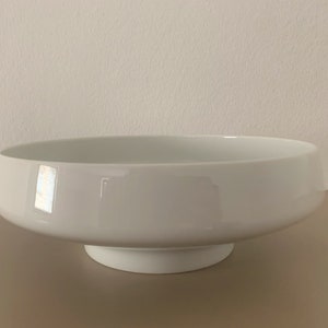 May include: A white ceramic bowl with a wide, shallow base and a raised lip. The bowl is sitting on a brown surface.