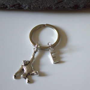Puede incluir: Llavero de plata con un pequeño amuleto de mono colgando de un anillo de plata con un amuleto de la letra E.