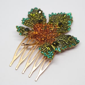 Op de afbeelding: Een gouden haar kam met een juweel-esdoornblad ontwerp. Het blad is gemaakt van groene, oranje en gouden strass stenen.