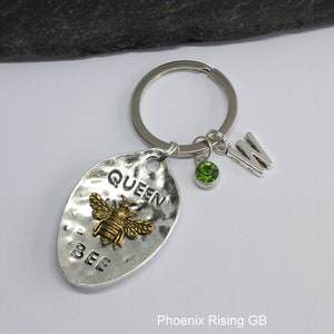 May include: Silver-coloured keyring with a hammered metal oval pendant. The pendant has the words "QUEEN BEE" and a gold-coloured bee. A green gemstone and the letter "W" dangle from the keyring.