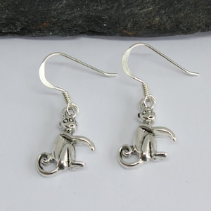 Può includere: Un paio di orecchini pendenti in argento a forma di scimmia. Le scimmie sono sedute con le braccia distese e le code arrotolate dietro di loro.