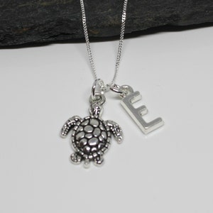 Könnte beinhalten: Eine silberne Kettenkette mit einem silbernen Schildkrötenanhänger und einem silbernen Buchstaben E-Anhänger.