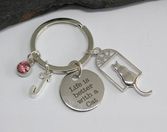 May include: Silver keychain with a charm that says "Life is better with a Cat" and a charm with a silhouette of a cat. The keychain also has a pink crystal charm and a silver letter J charm.