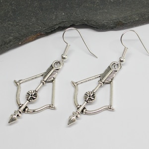 May include: Silver tone earrings featuring a bow and arrow design. The earrings are dangle style with a hook fastening.