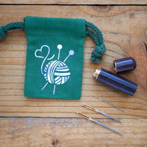 May include: A dark green drawstring pouch featuring a silver graphic of a yarn ball with knitting needles and a heart. Beside the pouch is a cylindrical dark wood needle case with a gold-colored metal cap and three silver sewing needles.