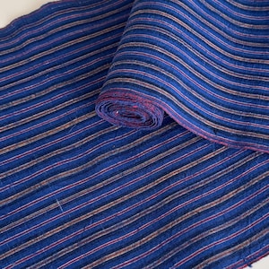May include: A roll of fabric featuring a deep blue base color with thin, repeating vertical stripes in shades of red, white, and black. The textile has a visible, slightly textured weave throughout, with the fabric partially unrolled to display.
