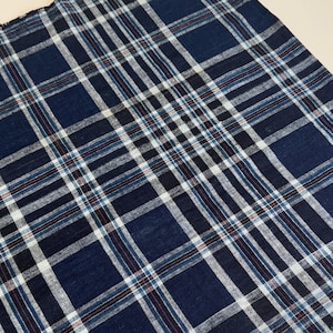 Op de afbeelding: Een stuk stof met een ruitpatroon in donker marineblauw, wit en zwart. Het textiel toont een raster van elkaar kruisende lijnen met dunne rode accenten door het hele ontwerp. De randen van het materiaal hebben een gerafelde, onafgewerkte textuur.