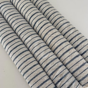 May include: Four cylindrical rolls of fabric arranged diagonally. Each roll features a natural beige base color with thin, evenly spaced dark navy blue horizontal stripes running across the entire length of the textured textile material.
