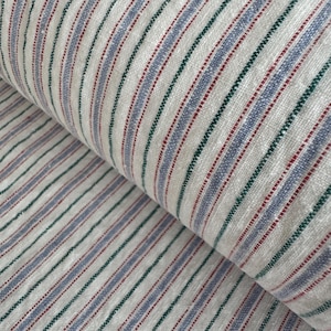 Può includere: Primo piano di un tessuto arrotolato con un motivo a righe. Il tessuto presenta strisce verticali alternate in bianco, blu, verde e rosso. Il tessuto sembra essere lino o un materiale simile tessuto.