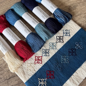 Könnte beinhalten: Sechs Spulen Sashiko-Garn in verschiedenen Blautönen, Rot und Weiß. Das Garn ist 100 Meter lang und wird in Japan hergestellt. Im Hintergrund befindet sich ein weißer Stoff mit blauen und roten Stickereien.
