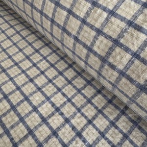 May include: A close-up of a rolled fabric featuring a blue and beige plaid design. The fabric has a textured surface, with blue lines creating a grid over the beige backdrop. The material appears to be linen or a similar textile.