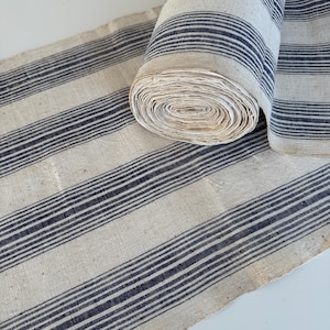 Puede incluir: Un rollo de tela color hueso con un patrón de rayas verticales finas de color azul oscuro. El tejido tiene una textura visible con pequeñas motas por todo el material, y el rollo está parcialmente desenrollado para mostrar el diseño a rayas.