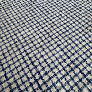 Op de afbeelding: Een geweven stof met een herhalend klein vichy-ruitpatroon in donker marineblauw en natuurlijk beige. Het textiel vertoont een zichtbare kruislings geweven textuur met een consistente rasterindeling over het gehele oppervlak van het materiaal.