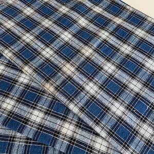 Op de afbeelding: Close-up van een blauw, wit en zwart geruite stof. De stof heeft een klassiek ruitpatroon met verschillende tinten blauw, wit en zwart. De stof is gevouwen, waardoor een gelaagd effect ontstaat. De stof lijkt katoen of een vergelijkbaar materiaal te zijn.