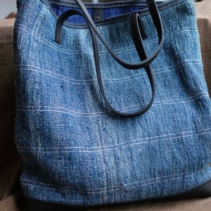 May include: A blue denim tote bag with black leather straps and a woven texture. The bag has a simple design and is made of durable material.