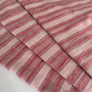 Peut inclure: Un tissu tissé présentant un motif répétitif de rayures verticales dans des tons de rose clair, de rouge sourd et de fines lignes grises. Le textile possède un tissage texturé visible avec une finition mate, plié en trois couches distinctes.