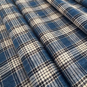 Op de afbeelding: Een geweven stof met een ruitpatroon in tinten blauw, wit en zwart. Het textiel vertoont een herhalend rasterontwerp met kruisende horizontale en verticale lijnen van variërende dikte, wat zorgt voor een getextureerd uiterlijk over het oppervlak.