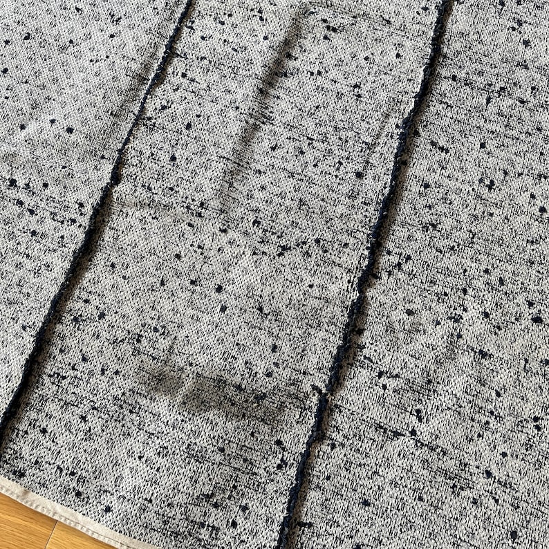May include: Close-up of a woven fabric with a white background and black speckles. The fabric has a textured, slightly rough surface.