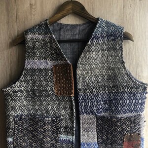 May include: A blue and grey patchwork vest with a brown leather patch and multiple pockets. The vest is made of a woven fabric and has a V-neck.