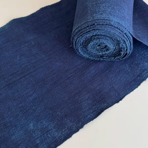 May include: A roll of indigo-dyed fabric with a textured weave. The fabric is a deep shade of blue, with variations in tone creating visual interest. The edge of the fabric is frayed, and the roll is partially unraveled.