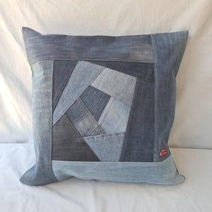 Könnte beinhalten: Quadratisches Denim-Kissen mit einem Patchwork-Design in verschiedenen Blautönen. Das geometrische Muster in der Mitte wird von einem dunkleren Denim-Rand umrahmt. Ein kleines rotes Etikett ist an der Seite sichtbar.