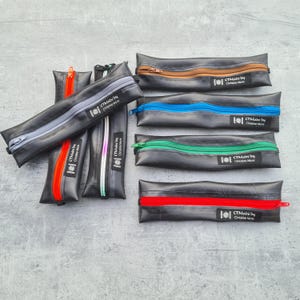 Könnte beinhalten: Eine Auswahl an schwarzen Federmäppchen mit farbenfrohen Reißverschlüssen in Rot, Orange, Blau, Grün und Silber. Jedes Etui hat eine rechteckige Form und einen Reißverschluss. Der Text "CTMade by Christine Muns" ist auf einem Etikett aufgedruckt.