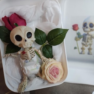 May include: A small skeleton figurine wearing a diaper, holding a light pink rose, and surrounded by red roses and green leaves. The figurine is in a white container with a blurred image of a similar skeleton holding a red rose.