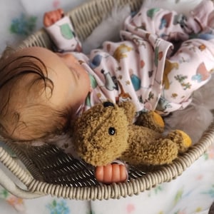 May include: A baby wearing a pink onesie with animal prints lies in a wicker basket with a brown teddy bear. The basket is lined with white fur and rests on a floral patterned blanket. The baby has light brown hair.