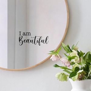 Peut inclure: Un miroir rond avec un cadre en bois affiche le texte "I am Beautiful". Un vase blanc contient un bouquet de fleurs. Une pile de livres et une tasse blanche avec une cuillère sont posés sur une surface en bois.