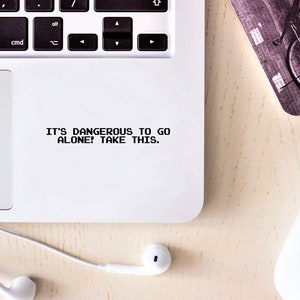 Op de afbeelding: Een witte laptop met een zwart toetsenbord en een trackpad. Het trackpad heeft een sticker met de tekst "IT'S DANGEROUS TO GO ALONE! TAKE THIS."