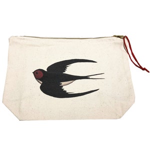 May include: A cream canvas zipped pouch with a black and white swallow bird illustration and a red head. It has a gold zip and a red cord pull. This cosmetic bag is ideal for storing makeup or other small items, and is a great gift.
