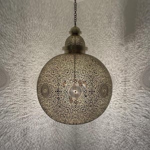 May include: A brass-colored, spherical pendant light fixture with intricate cut-out patterns. The light is suspended from a chain and ceiling mount. The design casts a decorative pattern of light and shadow on the surrounding walls.
