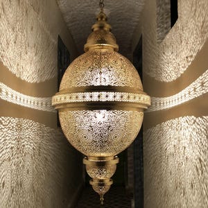 May include: A gold-colored, ornate pendant lamp hangs in a narrow hallway. The lamp has a spherical body with intricate cut-out patterns, casting decorative light and shadow patterns on the walls. The hallway has a patterned tile floor.