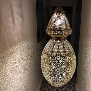 May include: Ornate, gold-colored pendant lamp with intricate cut-out patterns, casting patterned light on the surrounding walls. The lamp hangs in a narrow hallway with a patterned tile floor. The lamp's design is reminiscent of Moroccan or Middle Eastern styles.