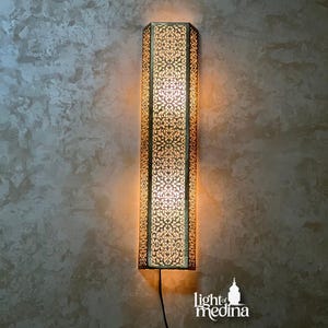 May include: A tall, vertical, hexagonal wall sconce featuring an intricate, laser-cut metal pattern that emits a warm, golden glow. The fixture has a metallic finish and includes a black power cord extending from the bottom. The text "light of medina" is visible in the lower right corner.