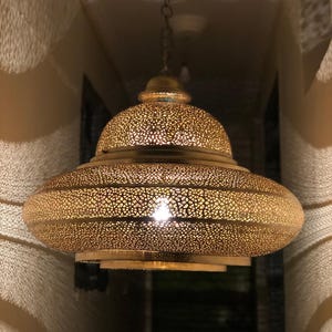 May include: A gold-colored, ornate pendant light fixture with a perforated design. The lamp has a domed top and a wide, horizontal band, suspended from a chain. The light shines through the intricate cutouts, creating a warm glow.