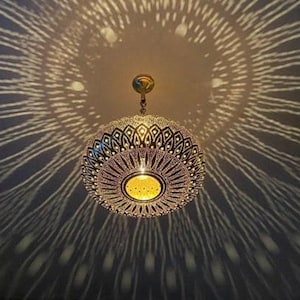 Puede incluir: Lámpara colgante esférica ornamentada con intrincados patrones calados. La luz proyecta un patrón radial en el techo, creando un efecto decorativo. La lámpara está suspendida de una cadena dorada.