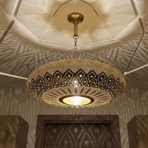 Pode incluir: Uma luminária pendente redonda de metal em tom de latão com padrões geométricos intrincados cortados a laser. A lâmpada é suspensa por uma corrente curta e uma haste central, projetando sombras simétricas detalhadas no teto acima.