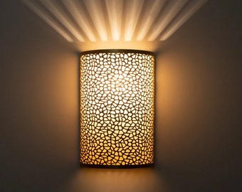 May include: A cylindrical wall sconce with a patterned design, emitting warm light. The sconce is made of a dark material, with a textured surface. Light radiates from the sconce, creating a fan-like pattern on the wall.