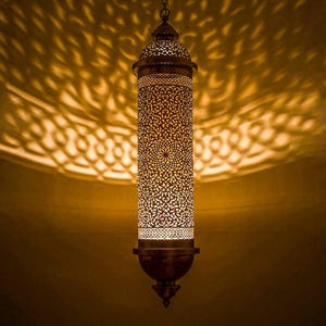 May include: A hanging, cylindrical pendant lamp with intricate, openwork metal detailing. The lamp is bronze-colored and emits a warm, inviting light, casting decorative shadows on the surrounding wall. The design features floral and geometric patterns.