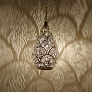 Op de afbeelding: Een decoratieve, druppelvormige hanglamp. De metalen kap heeft een patroon van kleine, gelijkmatig verdeelde gaten, die een patroonlicht op de muur werpen. De armatuur hangt aan een ketting.